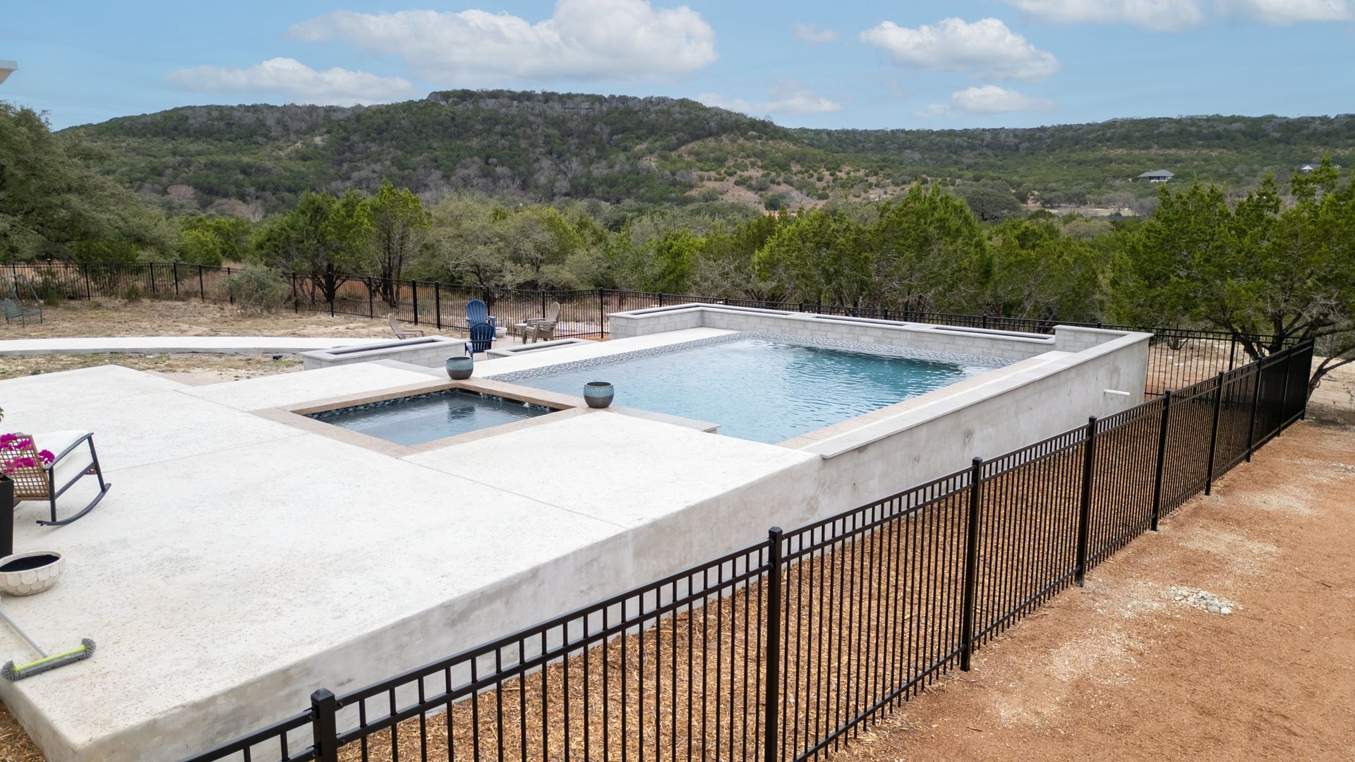 An elevated rectangular swimming pool and small spa on a light concrete patio, surrounded by a black fence and trees.