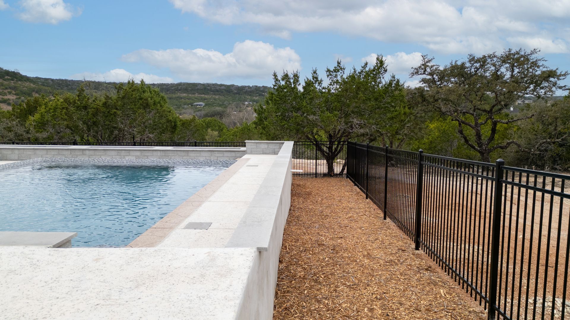 An infinity-edge swimming pool bordered by a stone ledge and black metal fence, overlooking a lush, hilly landscape.