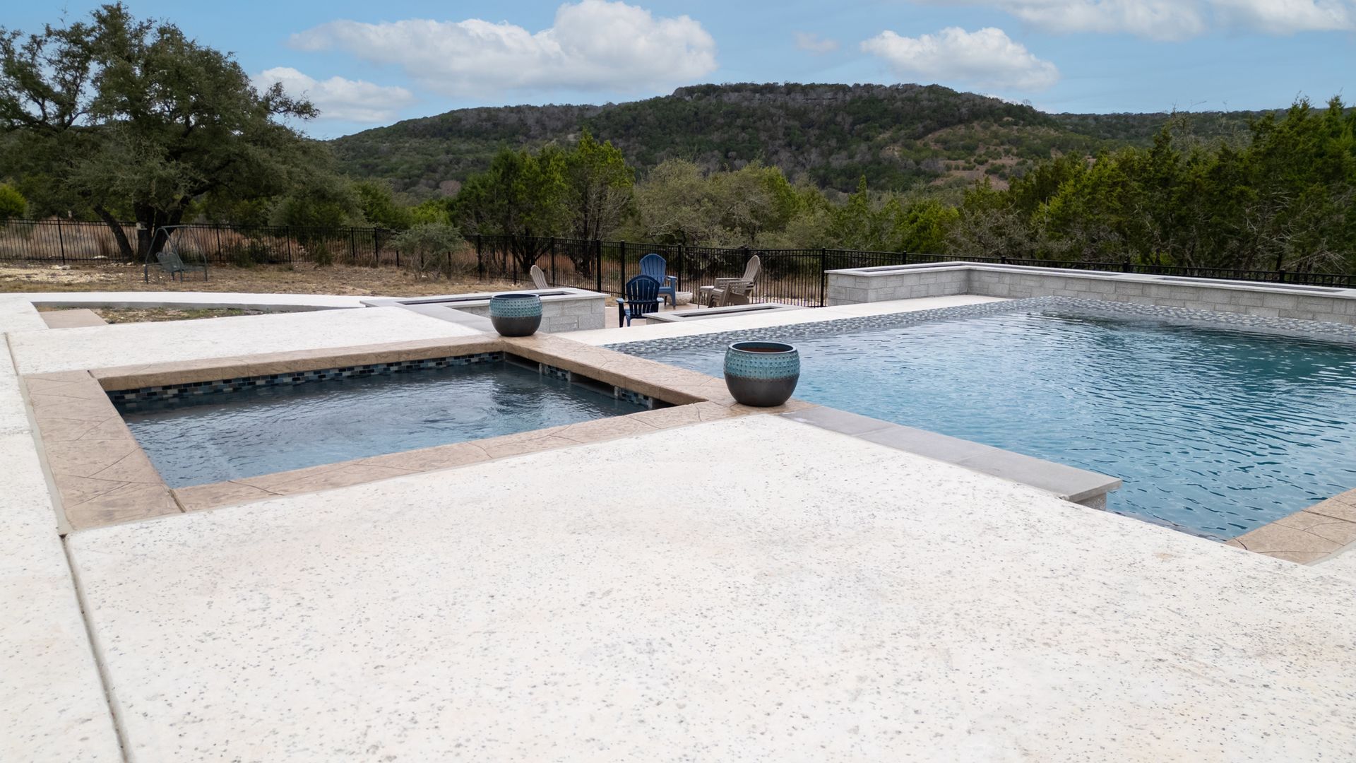 An outdoor swimming pool and spa with white concrete decking against a backdrop of trees and a hill under a blue sky.