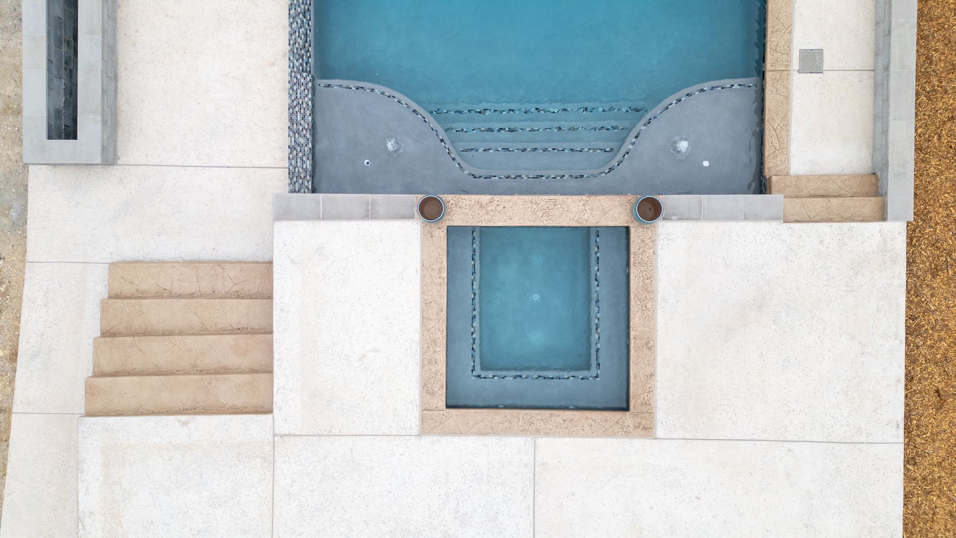 A top-down view of a rectangular swimming pool with a matching spa, surrounded by light-colored stone paving and steps.