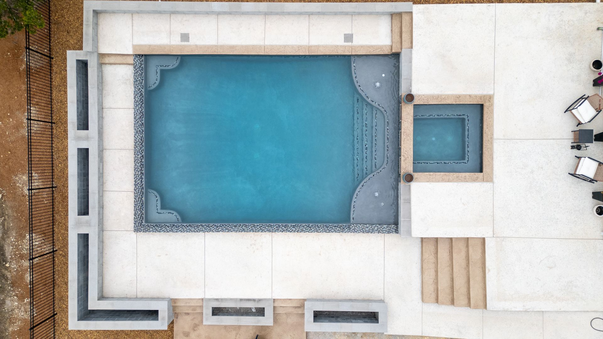 An aerial view of a rectangular blue swimming pool with an attached spa, surrounded by a light-colored concrete patio.