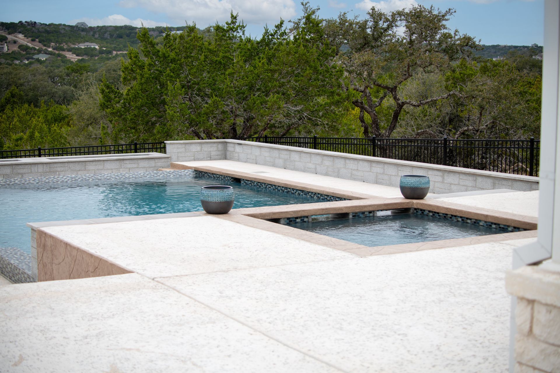 A swimming pool and attached hot tub with a light stone deck, set against a backdrop of green trees and hills.