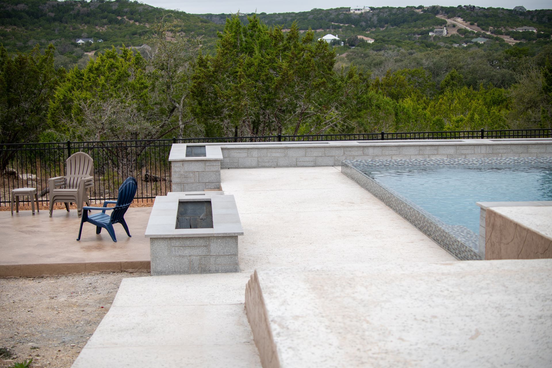 A stone pool deck and fire pit overlooking a wooded hillside, featuring two chairs near a backyard swimming pool.