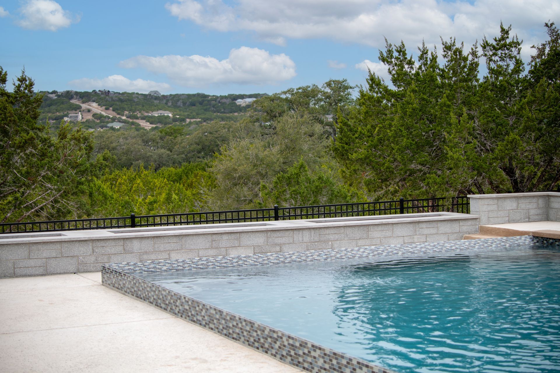 A turquoise swimming pool with a stone border overlooks a lush green valley under a bright, cloudy blue sky.