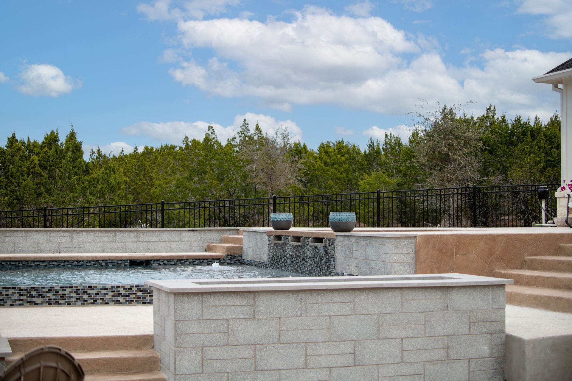 A modern backyard patio features a swimming pool with a waterfall, stone retaining walls, and a black fence by trees.