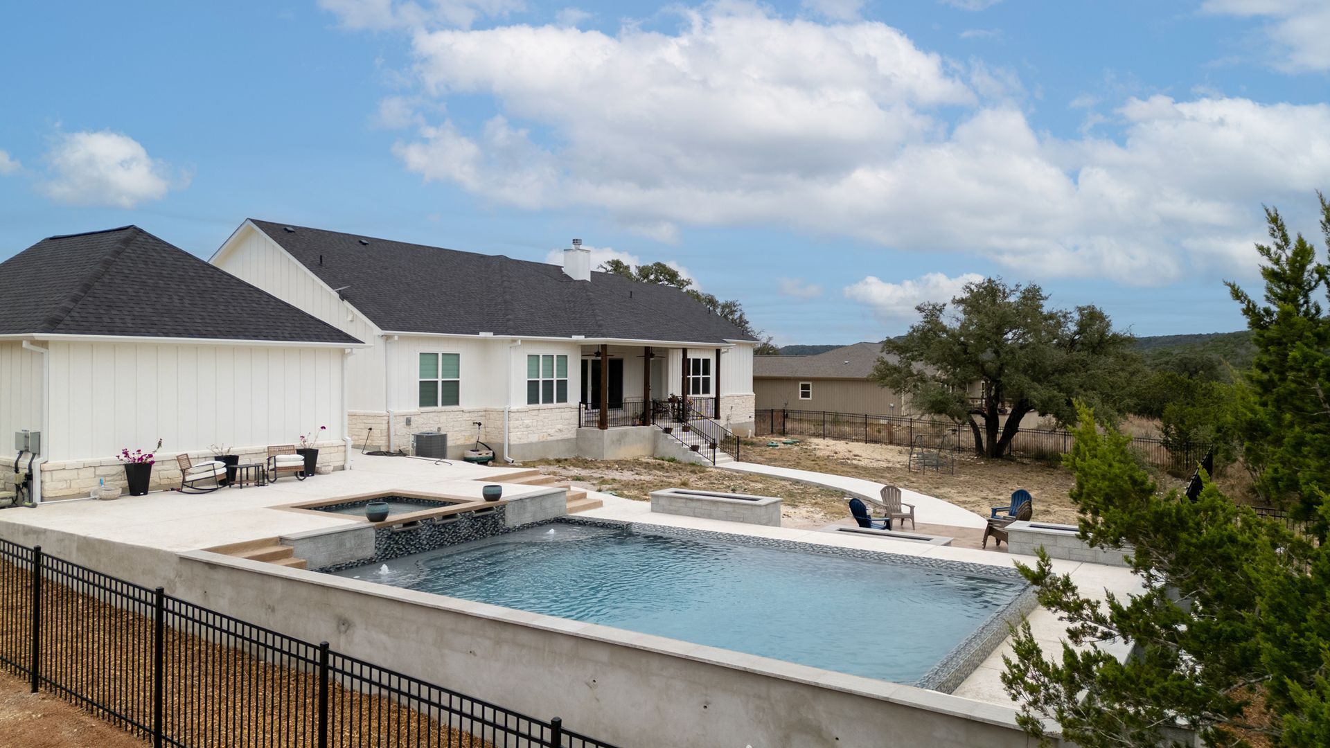 A light-colored house with a dark roof, a pool, and an attached hot tub in a backyard with a metal fence.