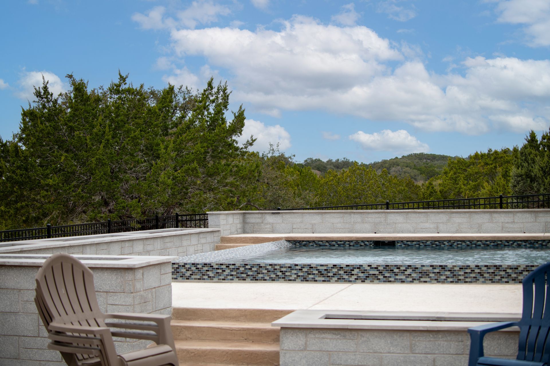 An infinity-style pool with a stone deck and two chairs, set against a backdrop of green trees and a blue sky with clouds.