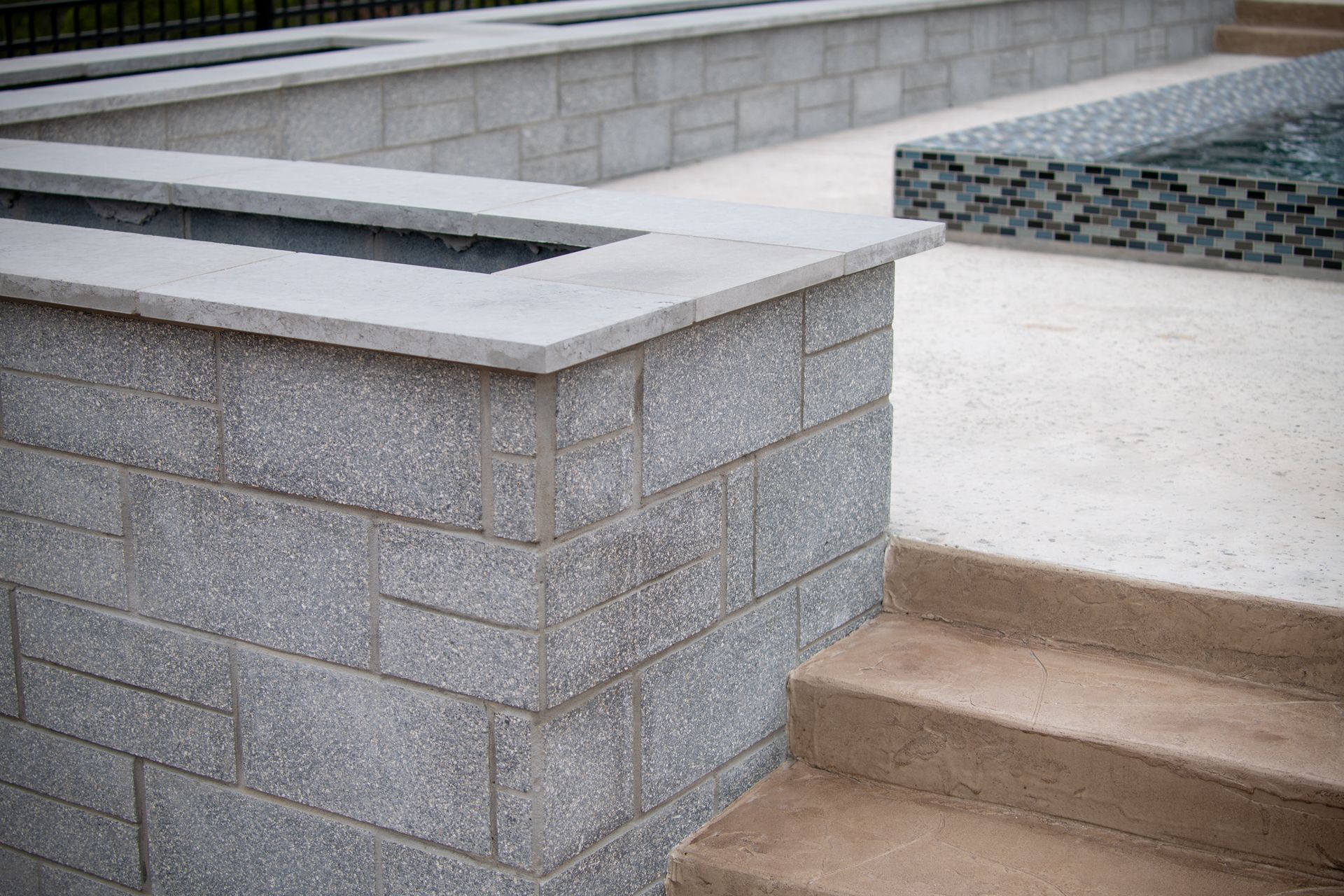 A gray stone retaining wall with a flat cap sits beside concrete stairs leading to a patio with a tiled water feature.