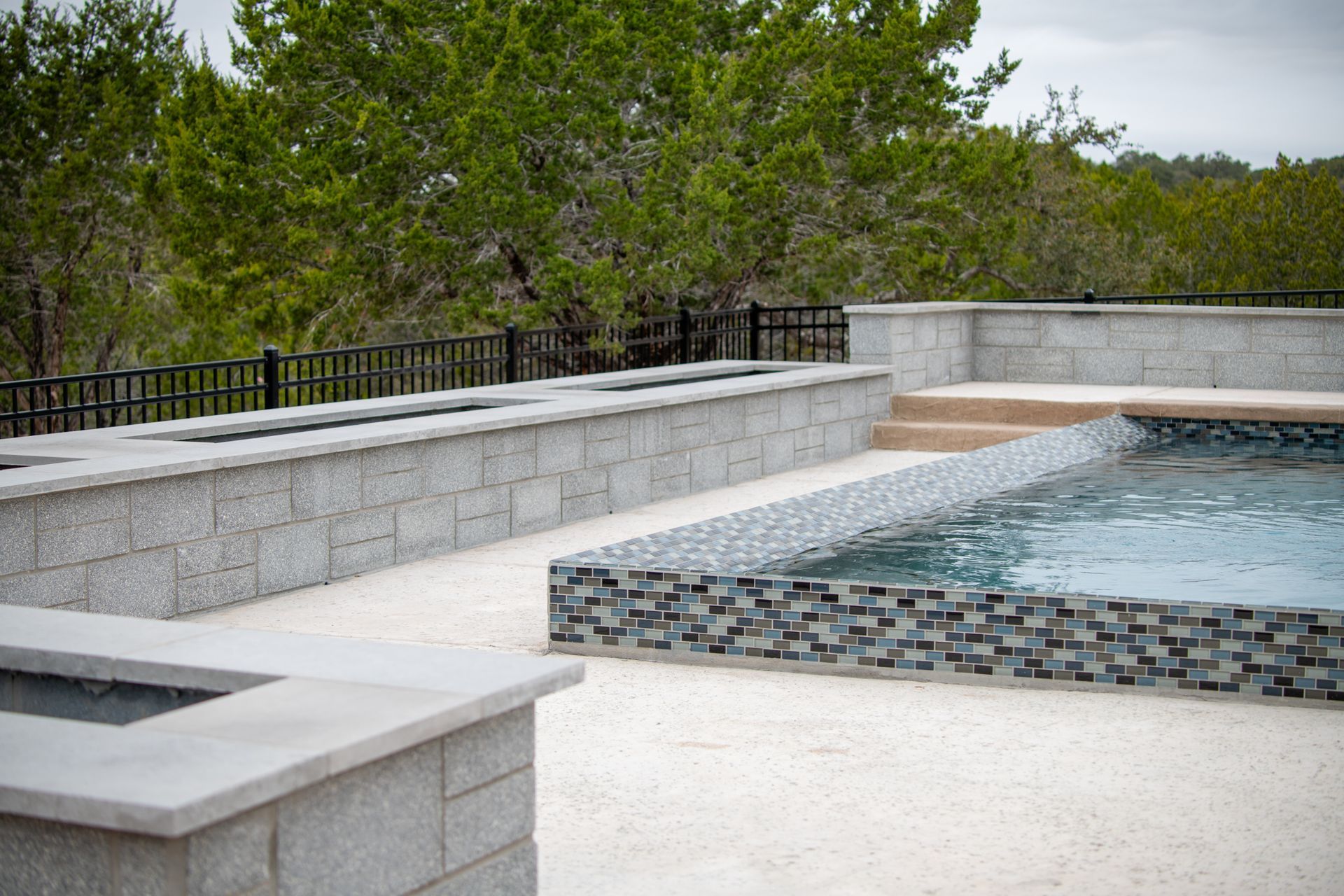 A patio features stone fire pits and a pool with decorative grey tile, bordered by a black fence and trees.