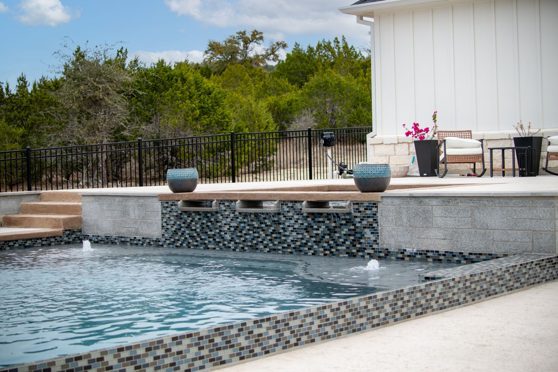 A swimming pool with three water features spilling from a gray stone wall, beside a patio with white siding and chairs.