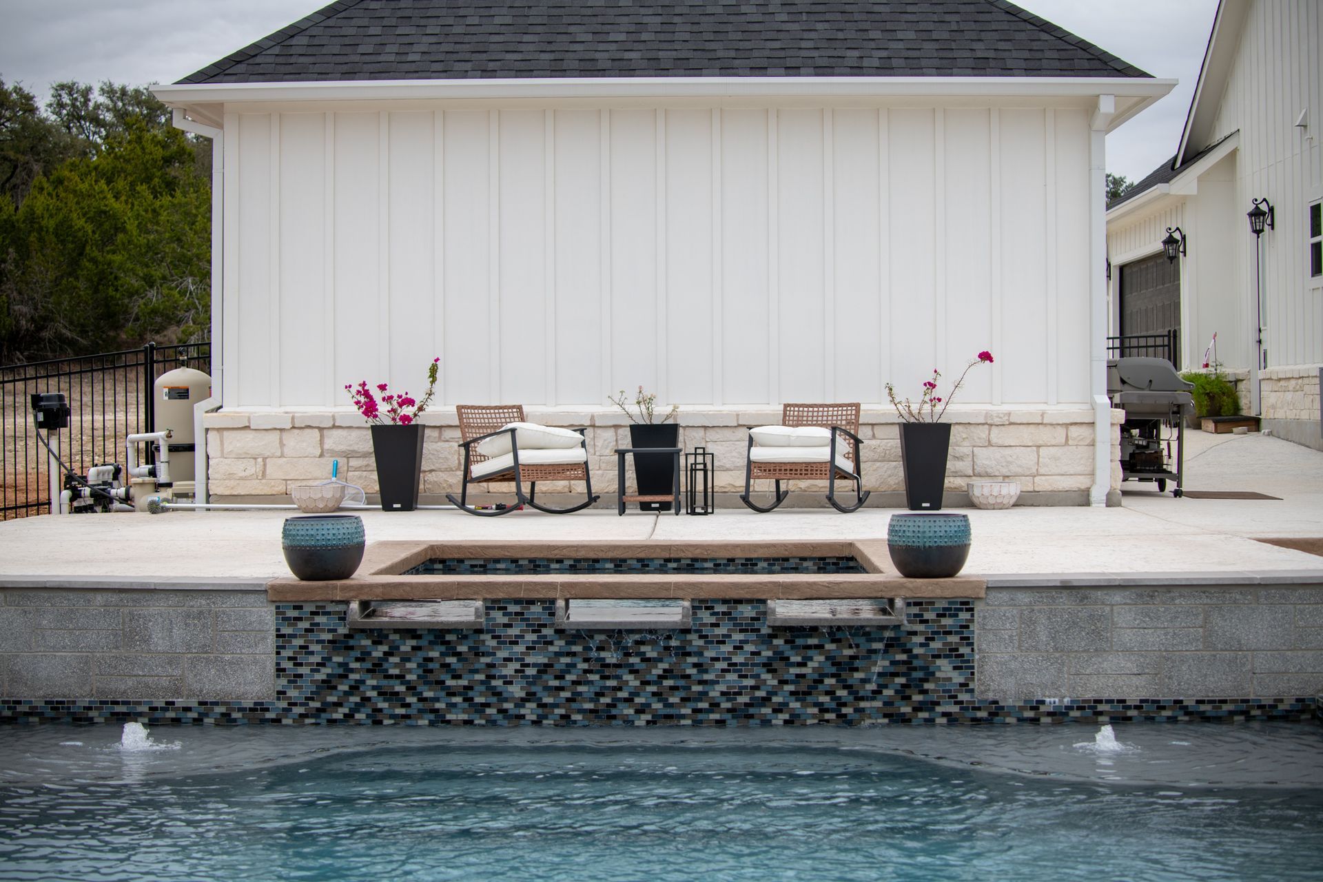 A poolside patio with two chairs, potted flowers, and a spa area in front of a white building with a dark roof.