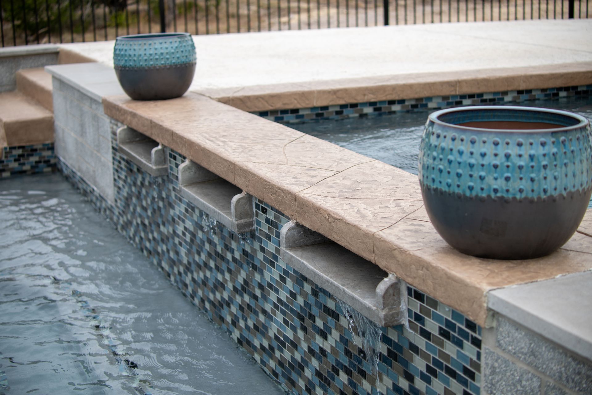 Blue and brown patterned pots sit on a tan stone ledge above a tiled spa with three cascading water features.