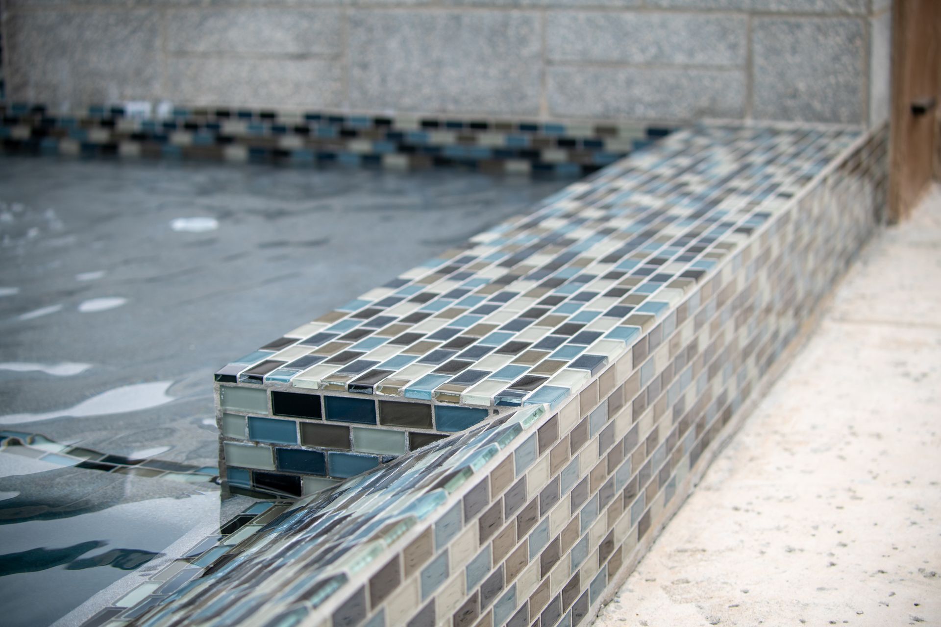 A swimming pool edge covered in small, rectangular glass tiles in varying shades of blue, grey, and brown.