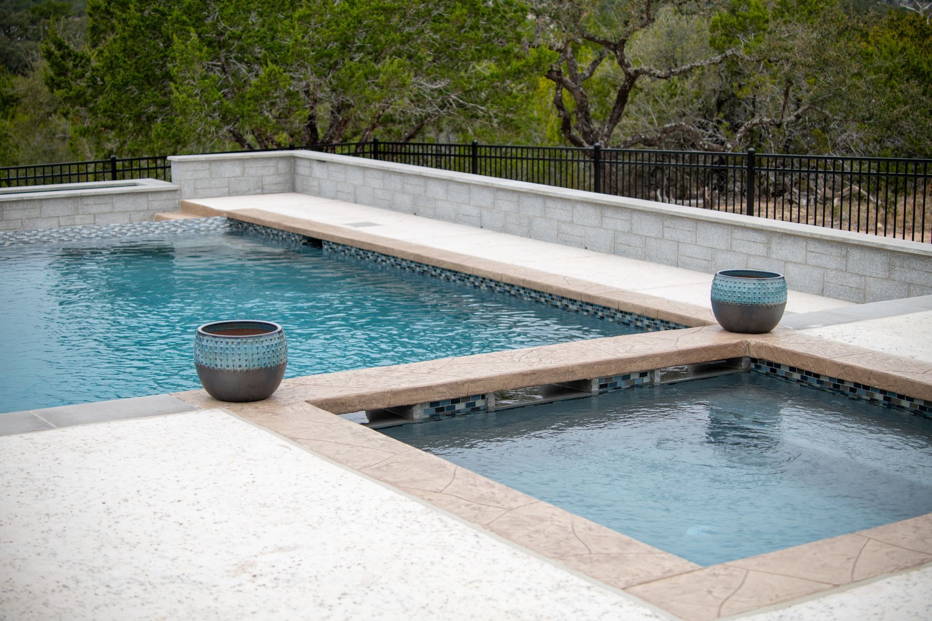A rectangular swimming pool and a separate square hot tub with light blue water, stone coping, and blue decorative pots.