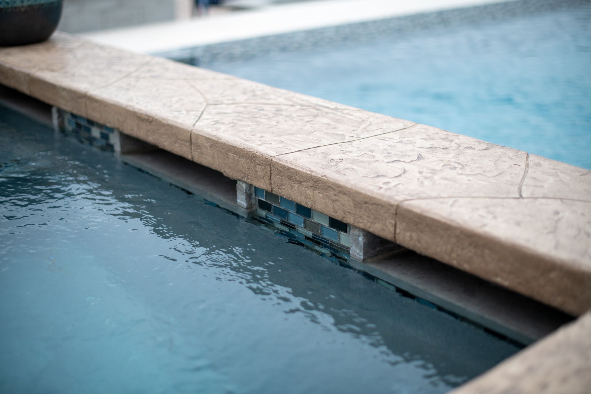 A close-up of a pool overflow edge with textured beige stone coping and blue mosaic tiles above the water line.