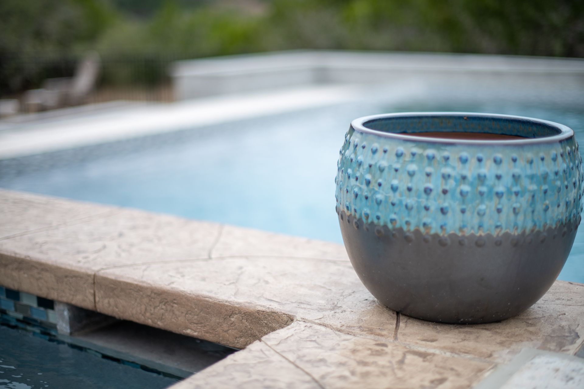 A two-toned, textured blue and dark grey pot sits on the stone edge of a swimming pool.