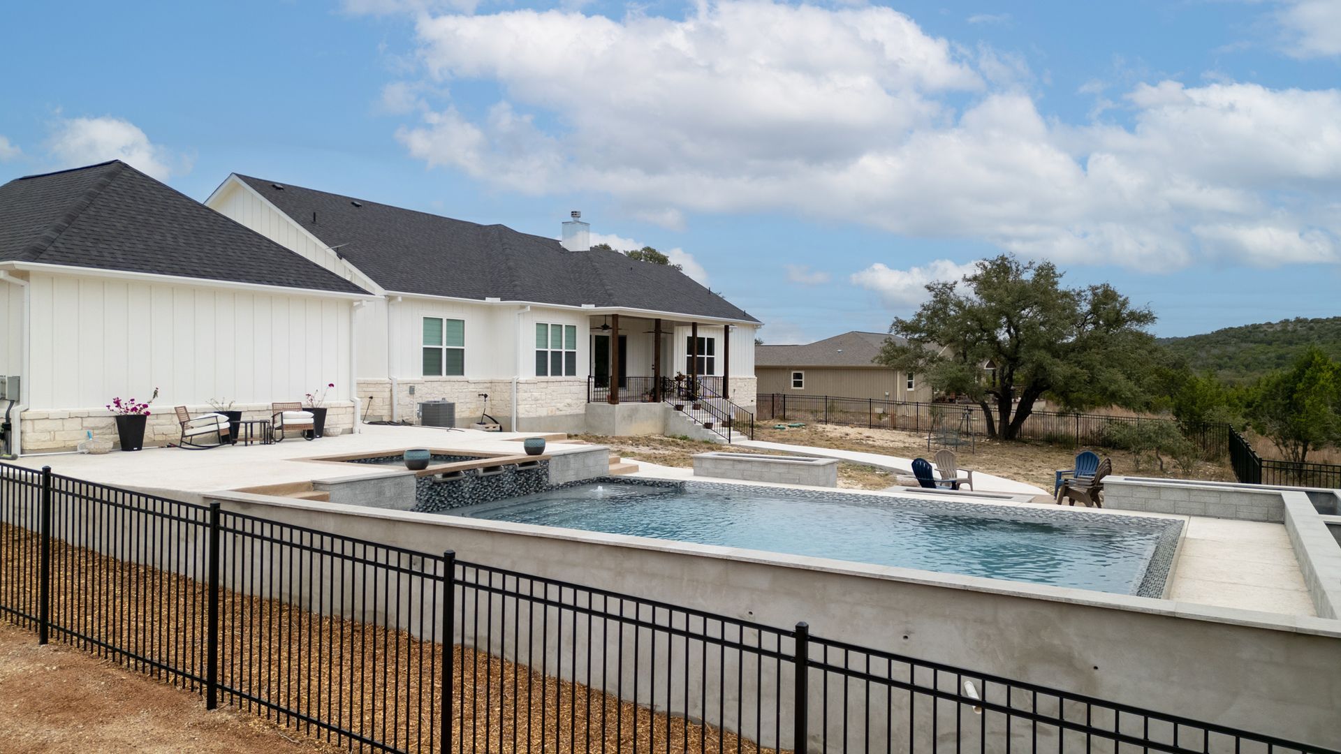 An elevated backyard pool with a water feature, set behind a black fence next to a white house under a cloudy blue sky.