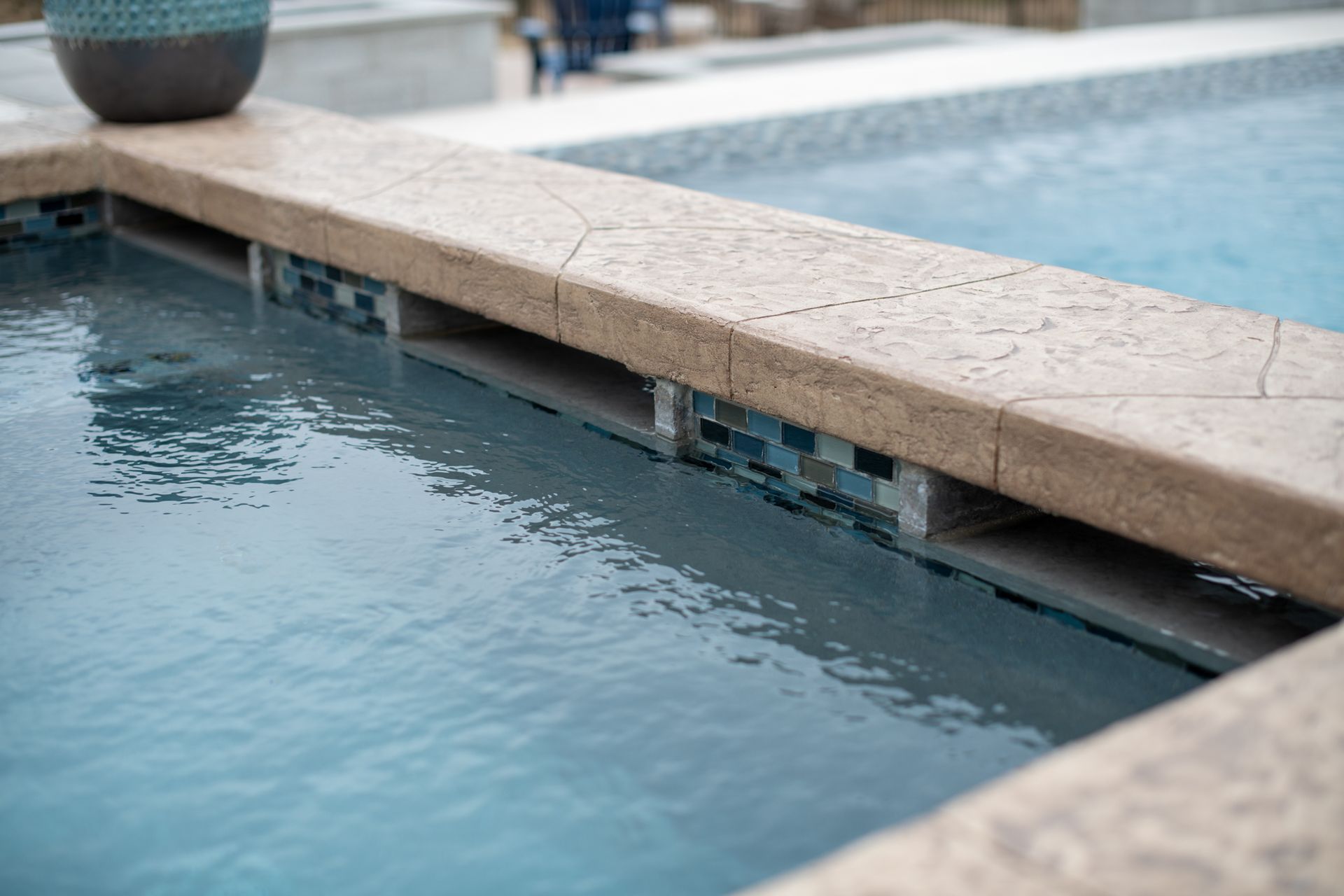 A close-up of a pool spa spillway with tan coping and blue mosaic tiles.