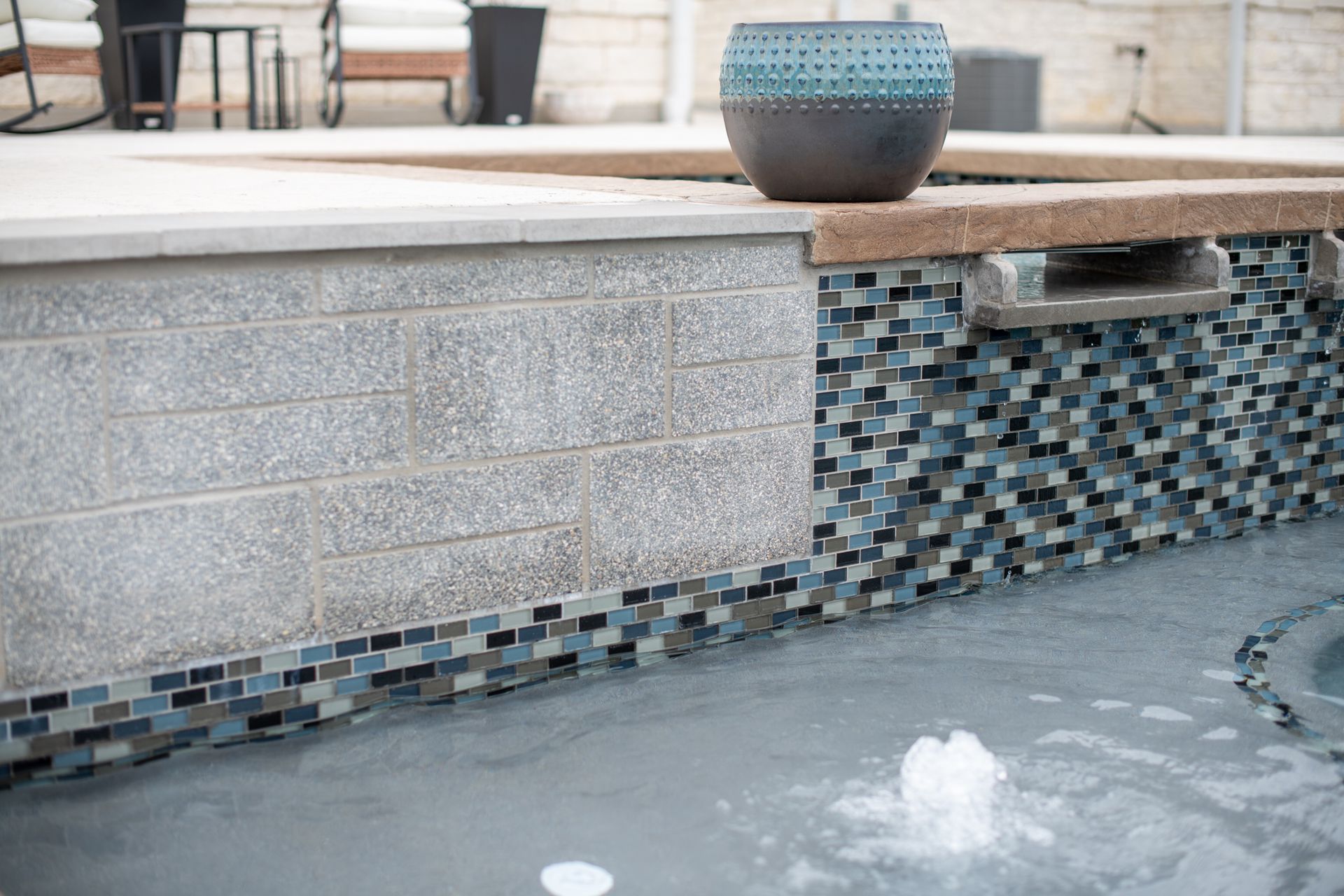 A pool water feature with a stone block wall, blue mosaic tile accents, and a small fountain bubbling in the water below.