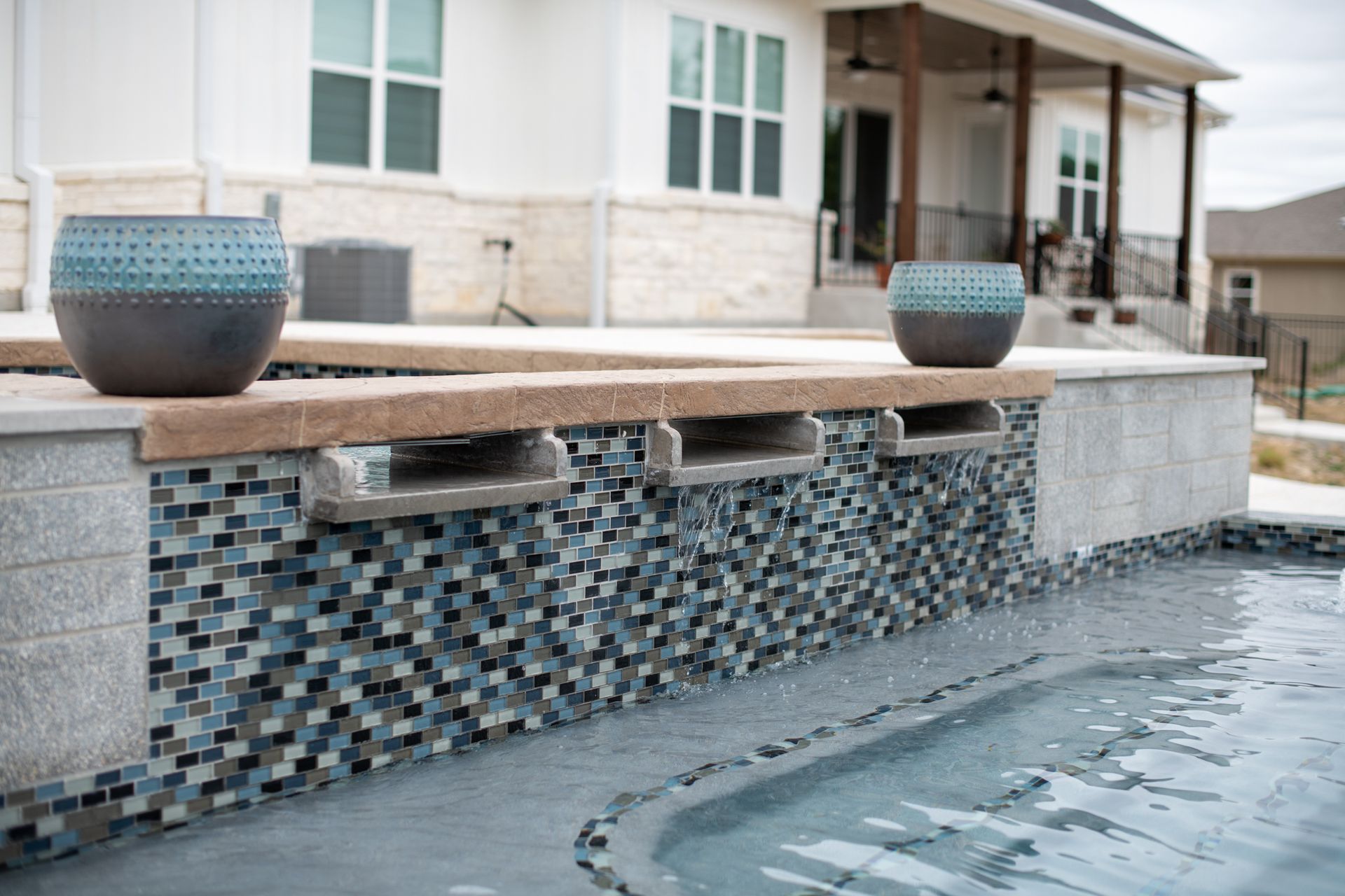 A pool spa with a tiled water feature, stone trim, and two decorative pots, with a house in the background.