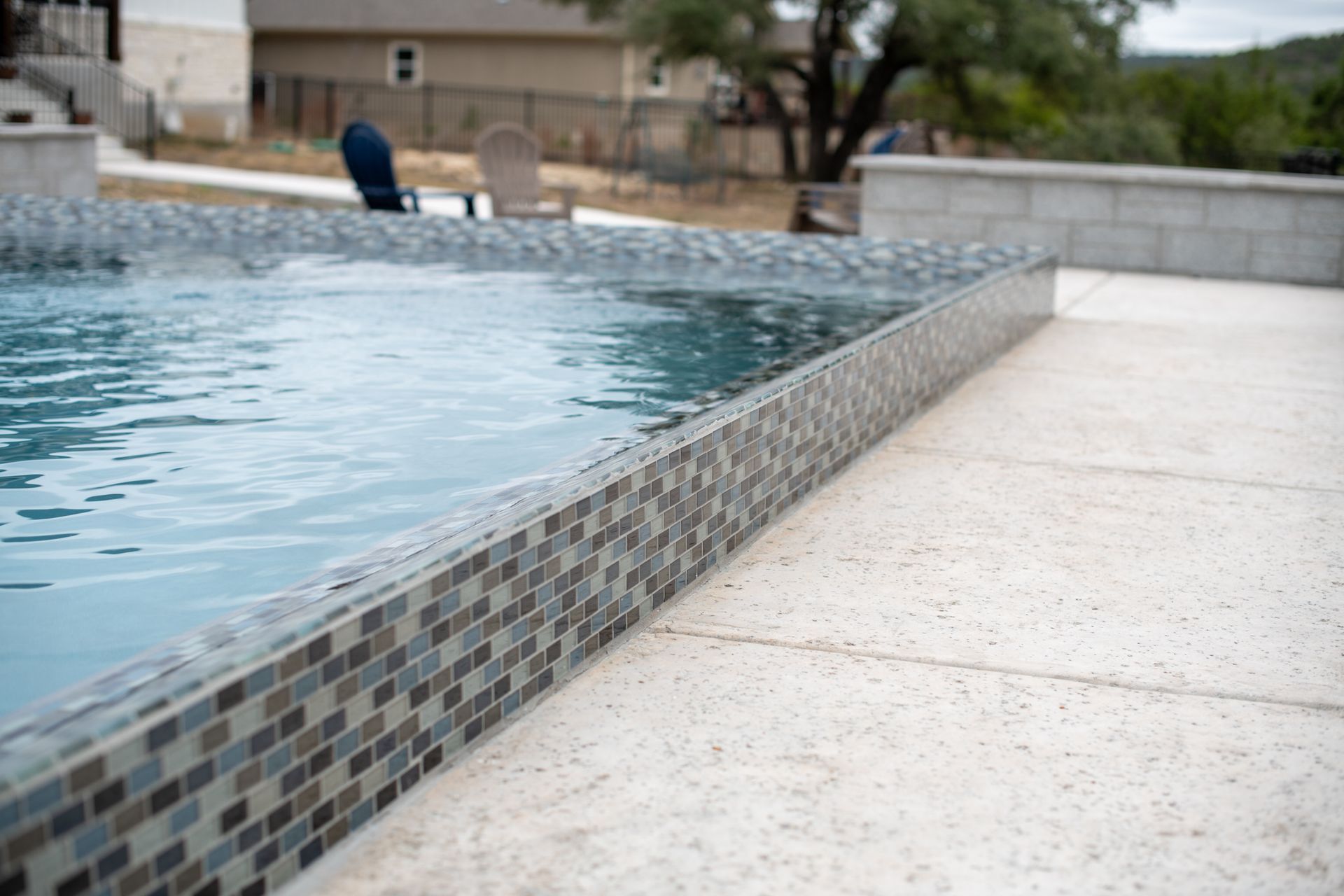 A close-up of a swimming pool edge with patterned mosaic tiles, next to a light-colored paved deck.