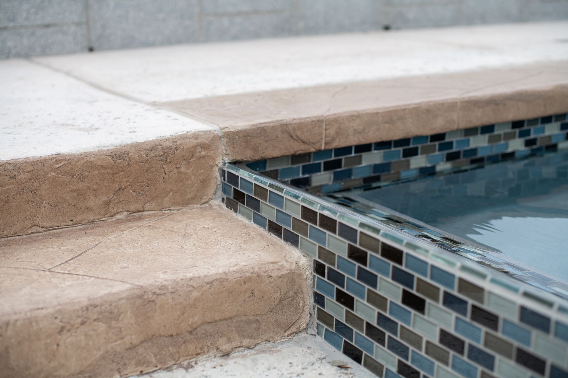 A close-up view of a pool edge featuring tan stone steps transitioning to a blue and gray tiled mosaic overflow wall.