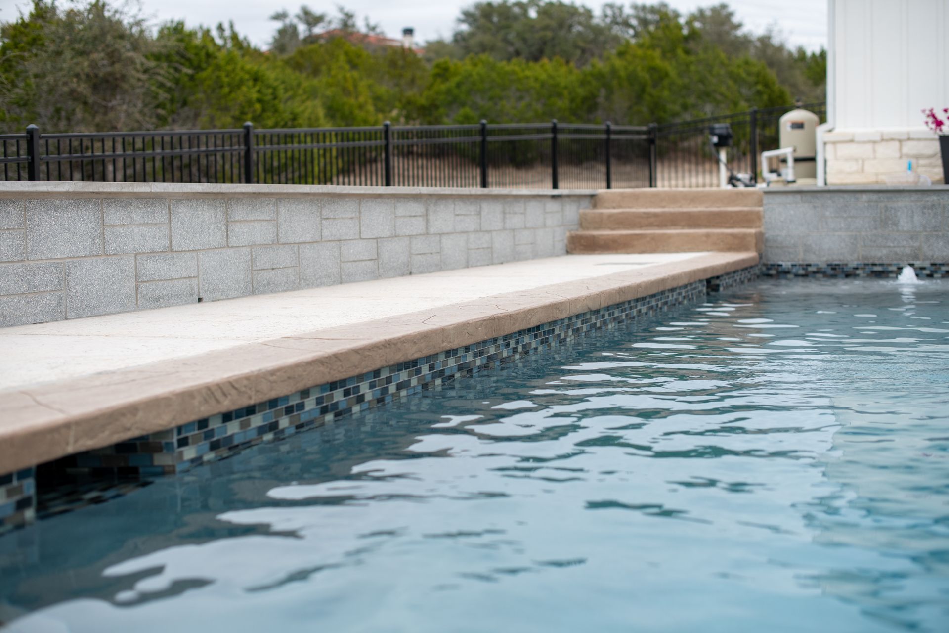 A swimming pool with blue water, a tiled edge, and concrete steps leading to a raised stone wall under a black fence.