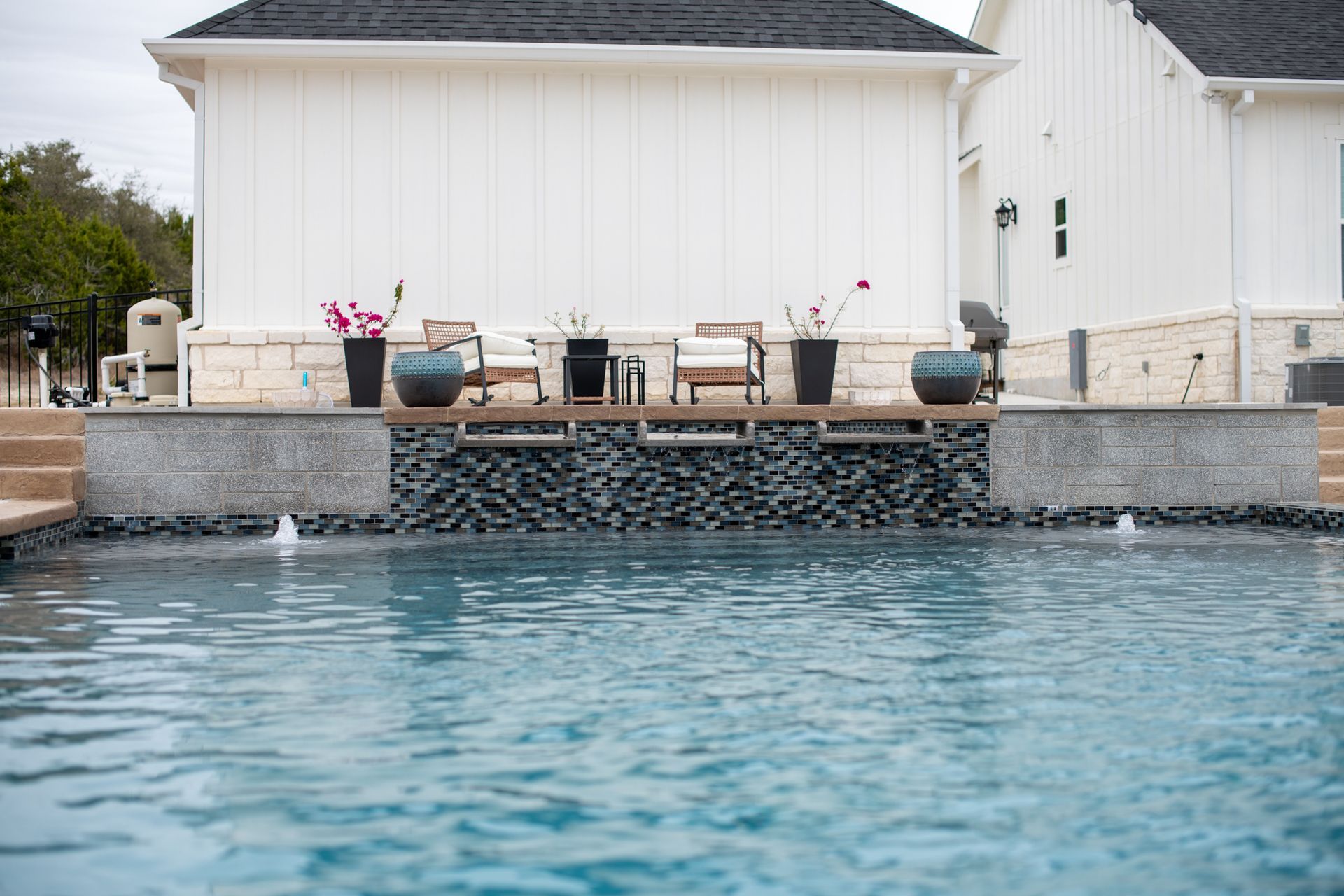 A swimming pool with two fountain jets in the foreground and a stone-walled patio with chairs and plants in the back.