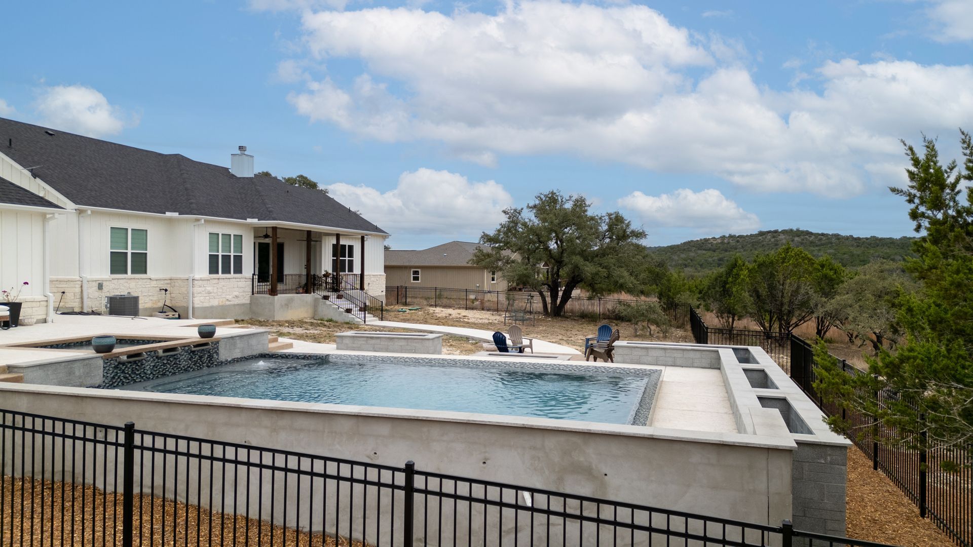 A modern white house with a dark roof overlooks a backyard swimming pool and a rural landscape under a cloudy blue sky.