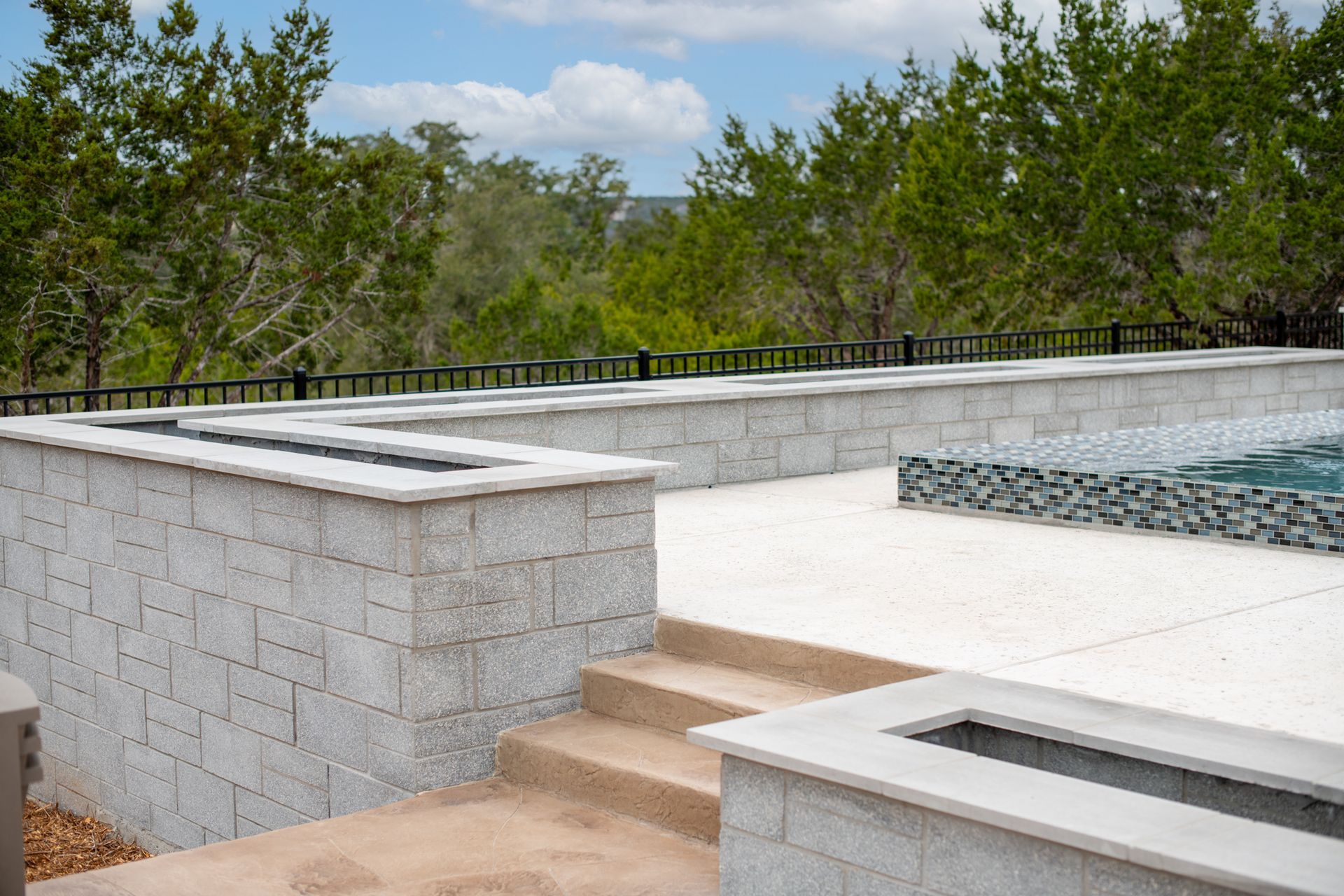 A modern light-colored stone patio with built-in fire pits, a swimming pool, and stone stairs overlooking a forest.