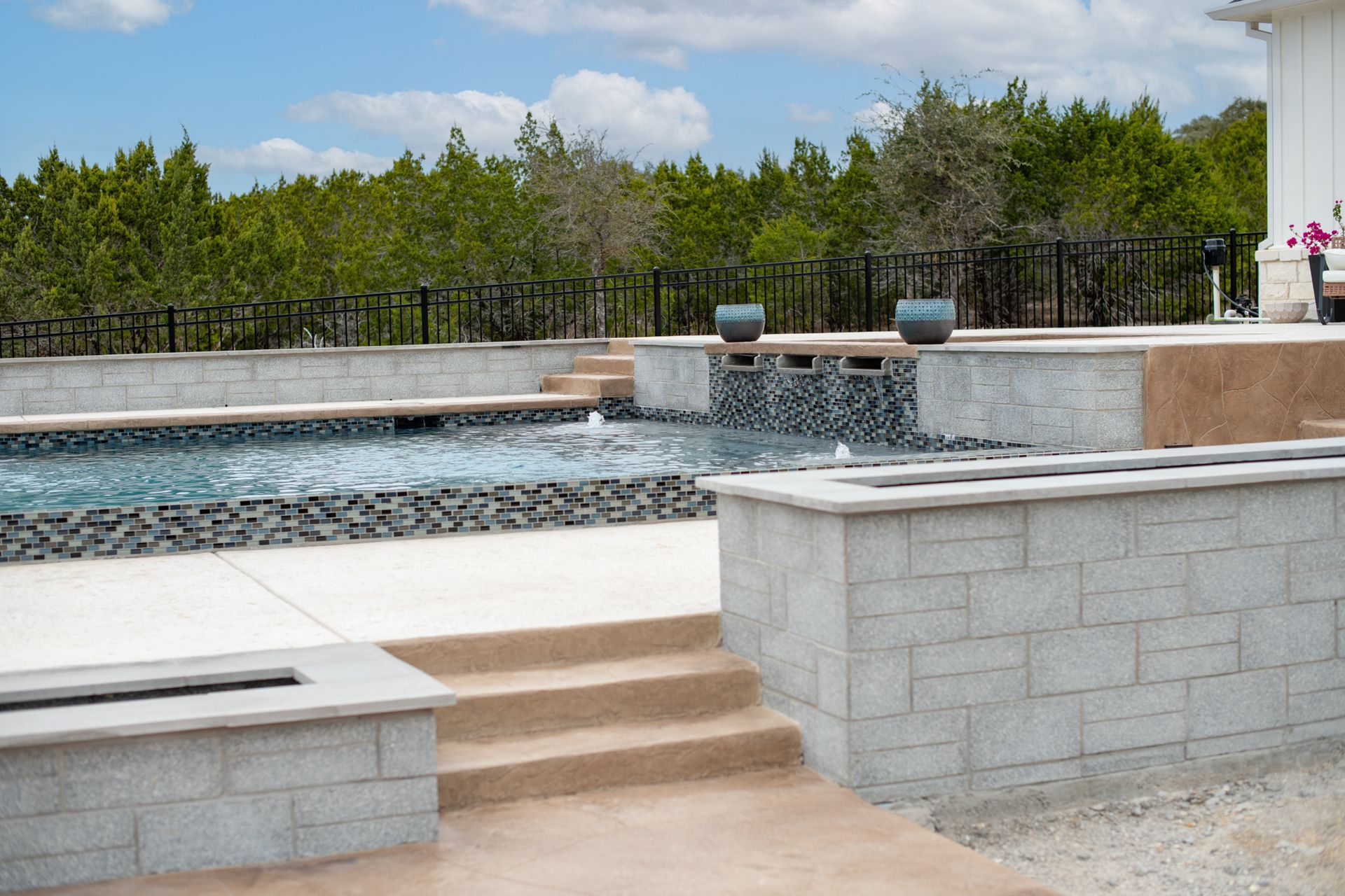 A modern backyard pool featuring stone steps, a cascading water feature, and a raised concrete patio under a blue sky.