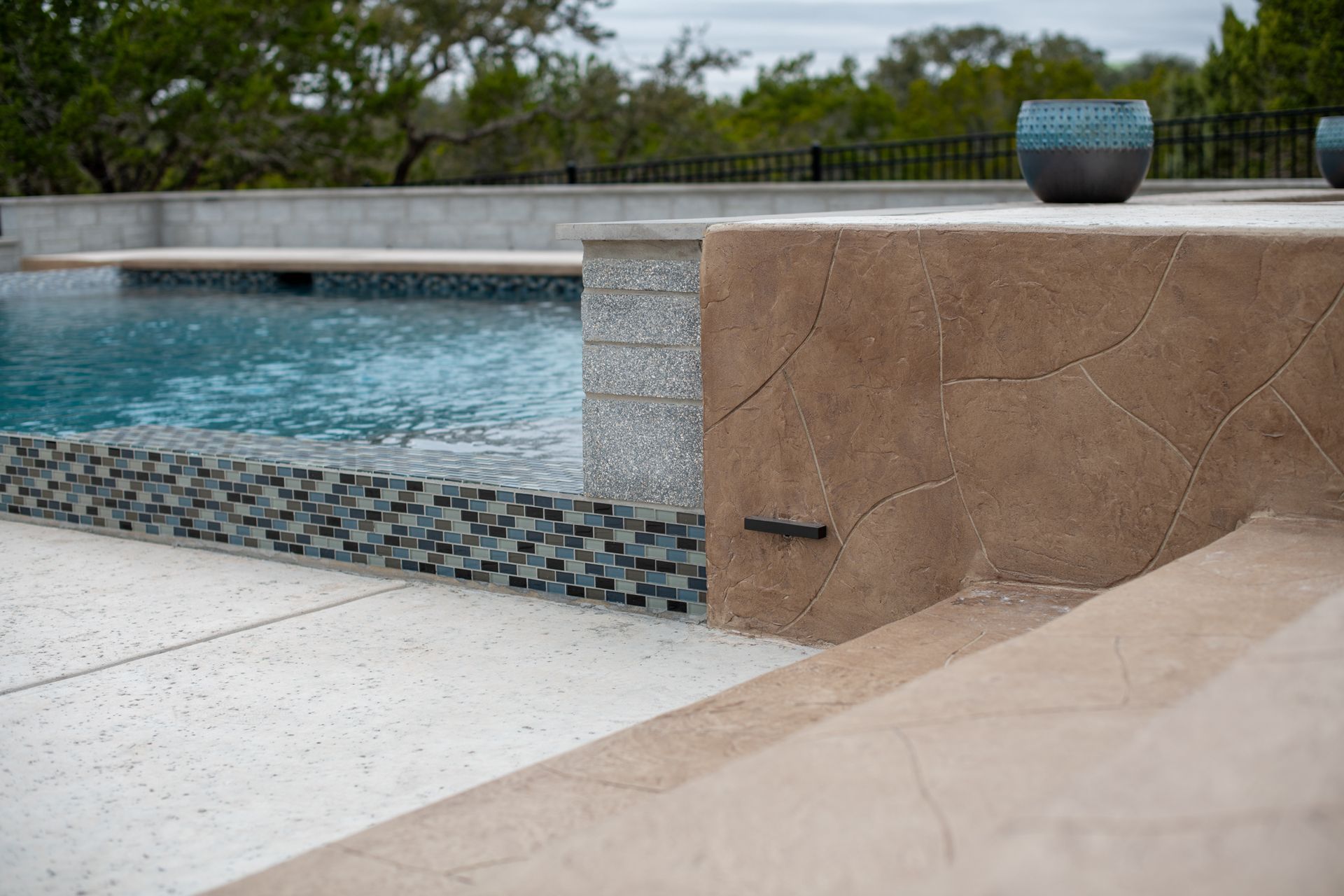 A concrete pool deck edge with tiled accents, a tan textured wall, and a view of the water and surrounding trees.