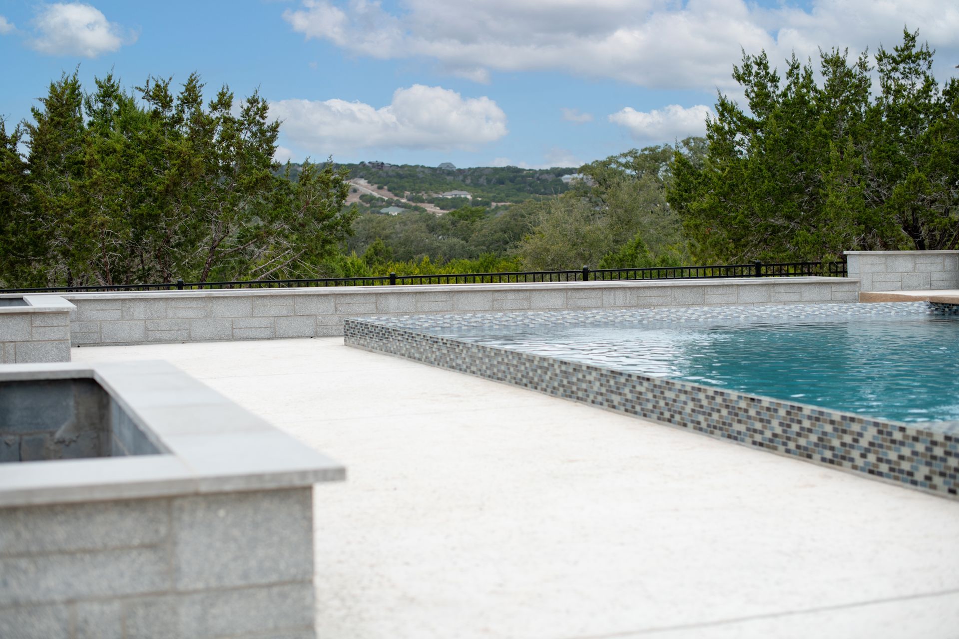 A swimming pool with mosaic tile edging sits on a stone patio overlooking a tree-filled hill landscape under a blue sky.