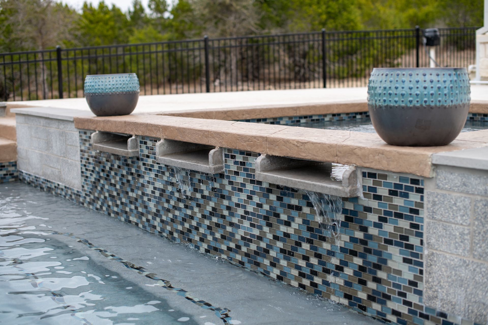 A pool wall with decorative blue and gray mosaic tiles, stone spillway fountains, and two matching patterned pots on top.