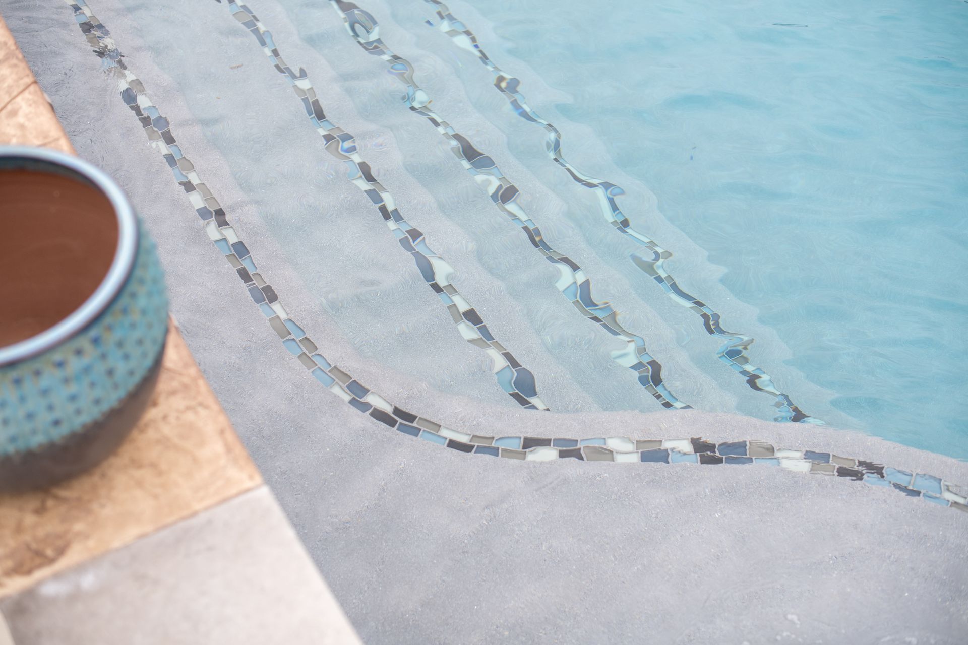 Top-down view of pool steps with decorative stone tile accents, next to a textured blue and brown pot on the deck.