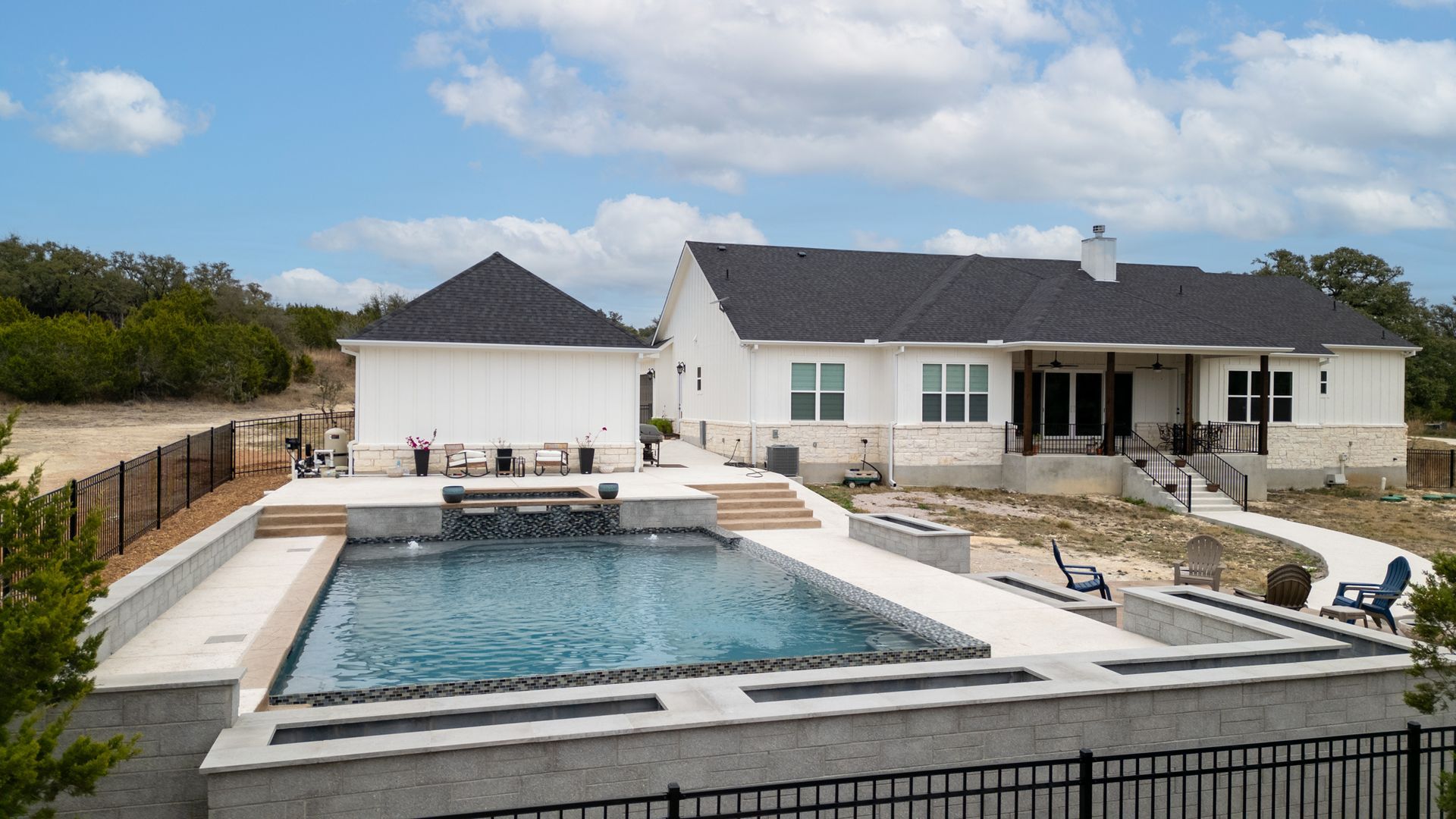 A modern white house with a dark roof overlooks a backyard pool with a built-in stone fire feature under a blue sky.