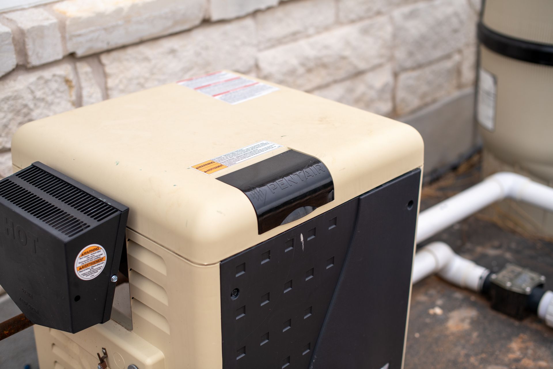 A beige pool heater with a black side panel and attached vent sits on a concrete patio next to white piping.