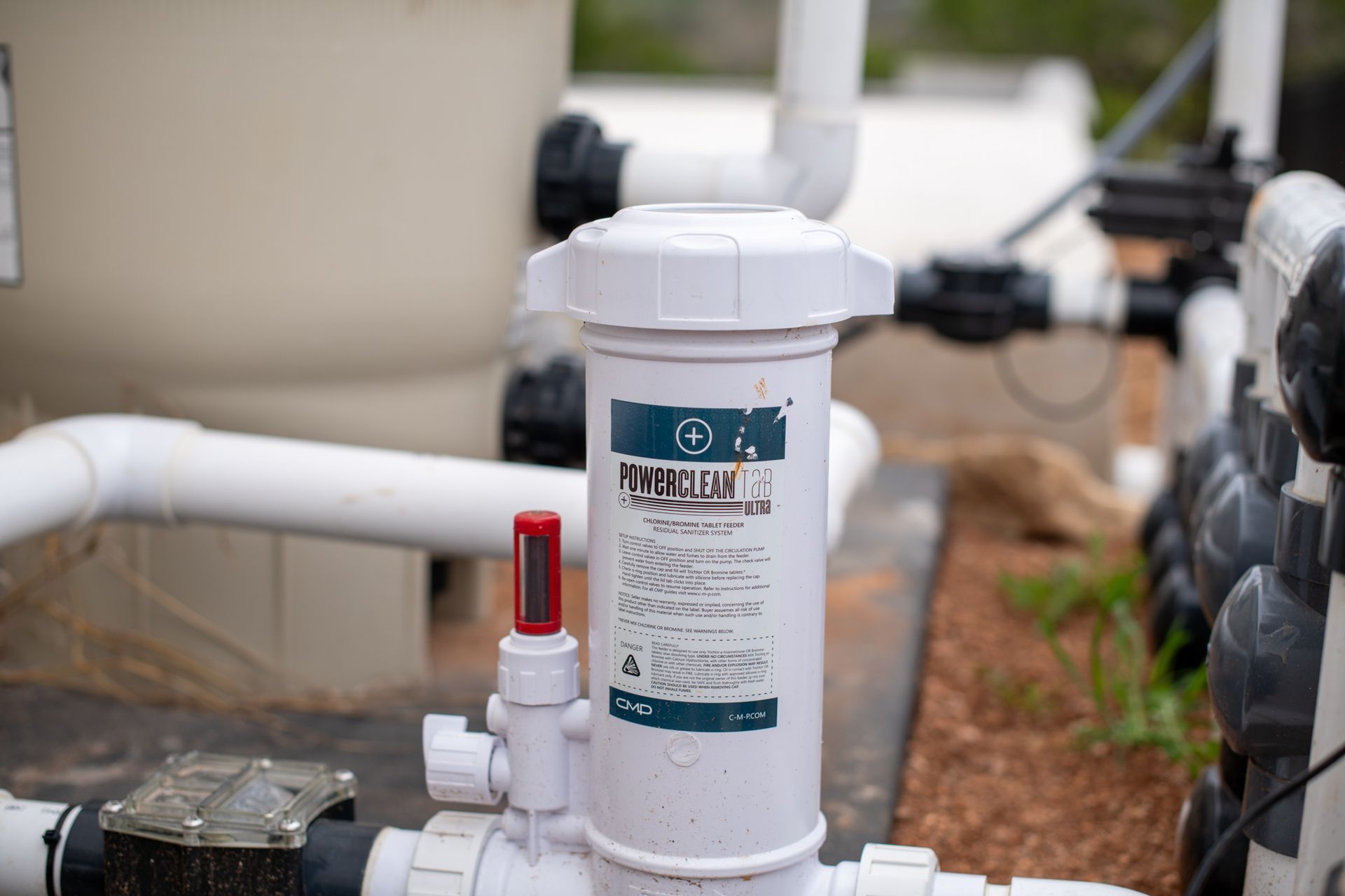 A white pool chemical feeder with a red flow gauge installed on plumbing near a large filter tank.