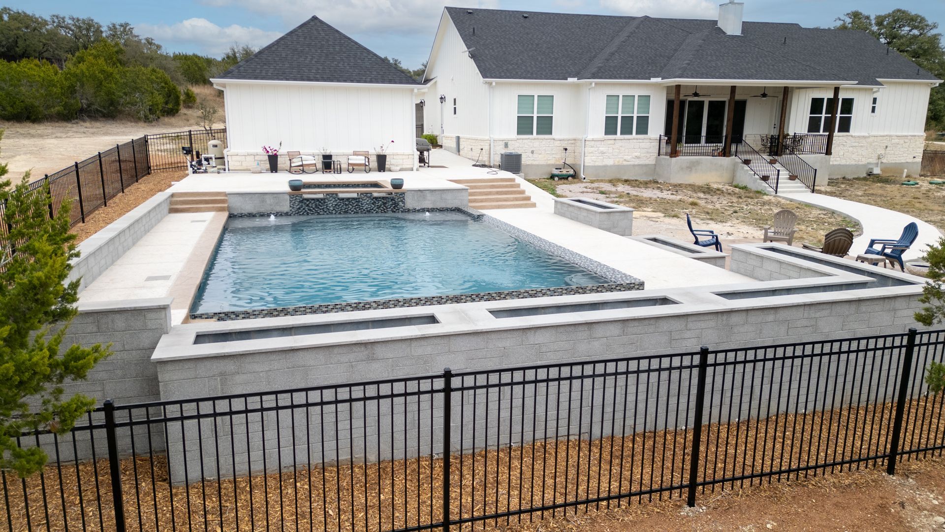 A modern rectangular pool with a stone wall, water feature, and patio, set behind a black fence in a suburban backyard.