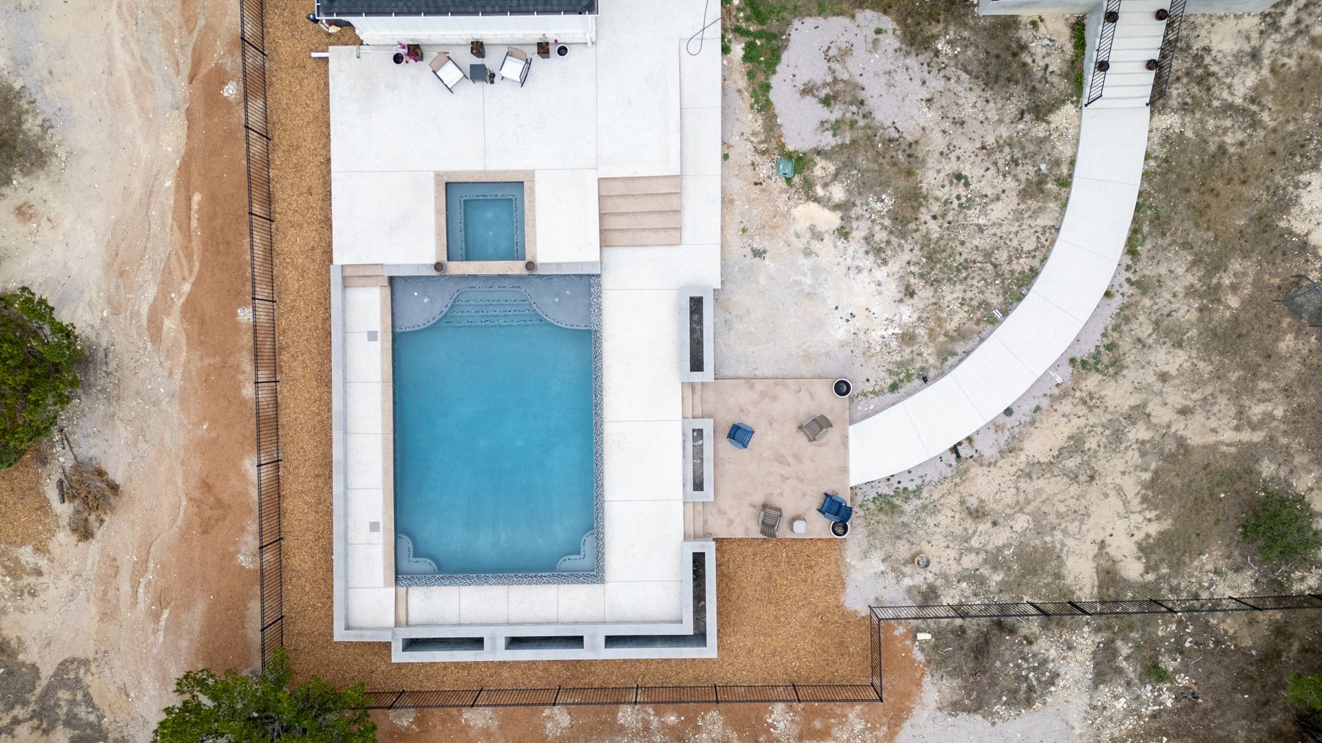 An aerial view of a backyard with a rectangular pool, a spa, concrete decking, and a curved walkway on dirt terrain.