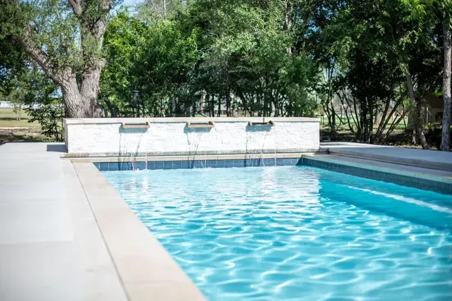 A rectangular backyard swimming pool featuring a white stone wall with three small water features flowing into the water.