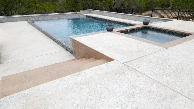 An elevated swimming pool and adjacent square hot tub set into a light-colored concrete deck with stone steps.
