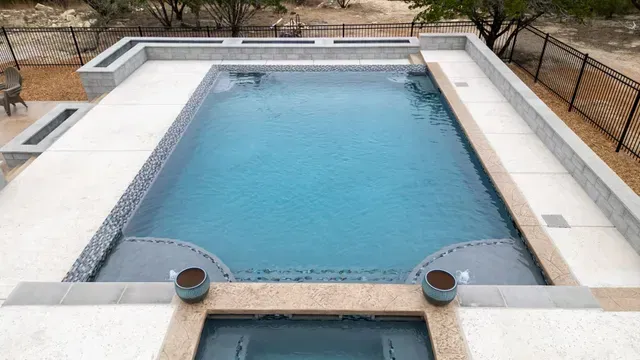 A rectangular blue swimming pool with a connected hot tub, surrounded by light-colored pavers and a metal fence.