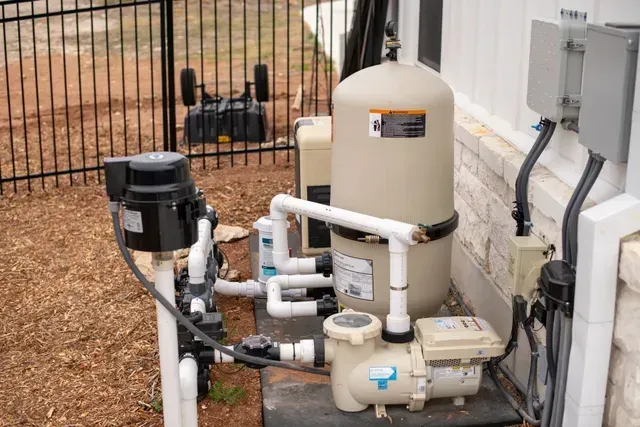 Pool pump and filter equipment installed on a concrete pad next to a stone wall and black metal fence.