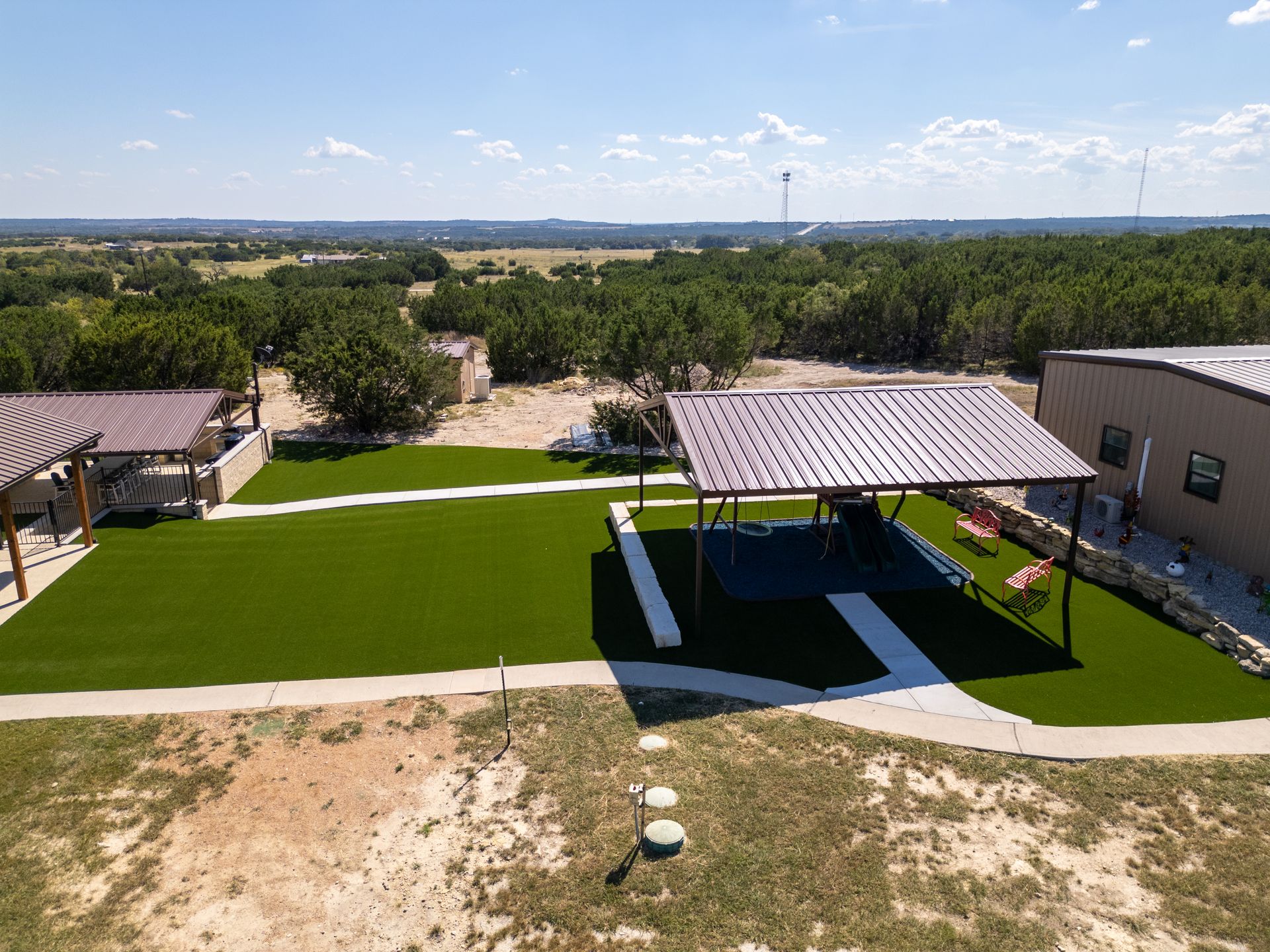 Aerial view of an artificial grass lawn with a metal-roofed pavilion and walkway between two buildings on a sunny day.