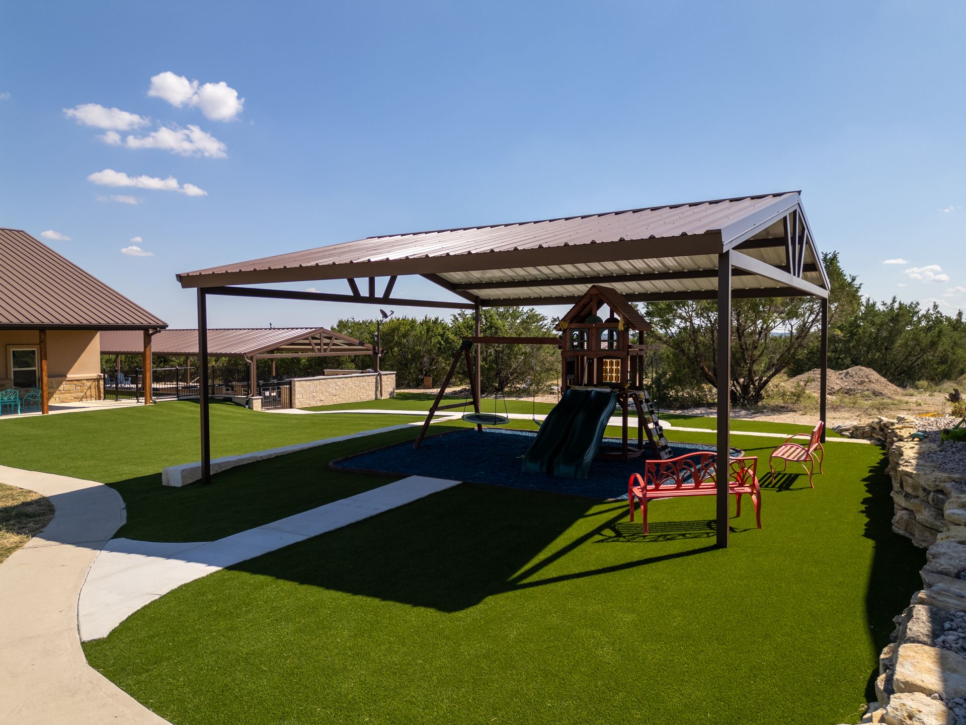 A playground with a slide and swing set under a large metal pavilion on a sunny day with artificial grass.