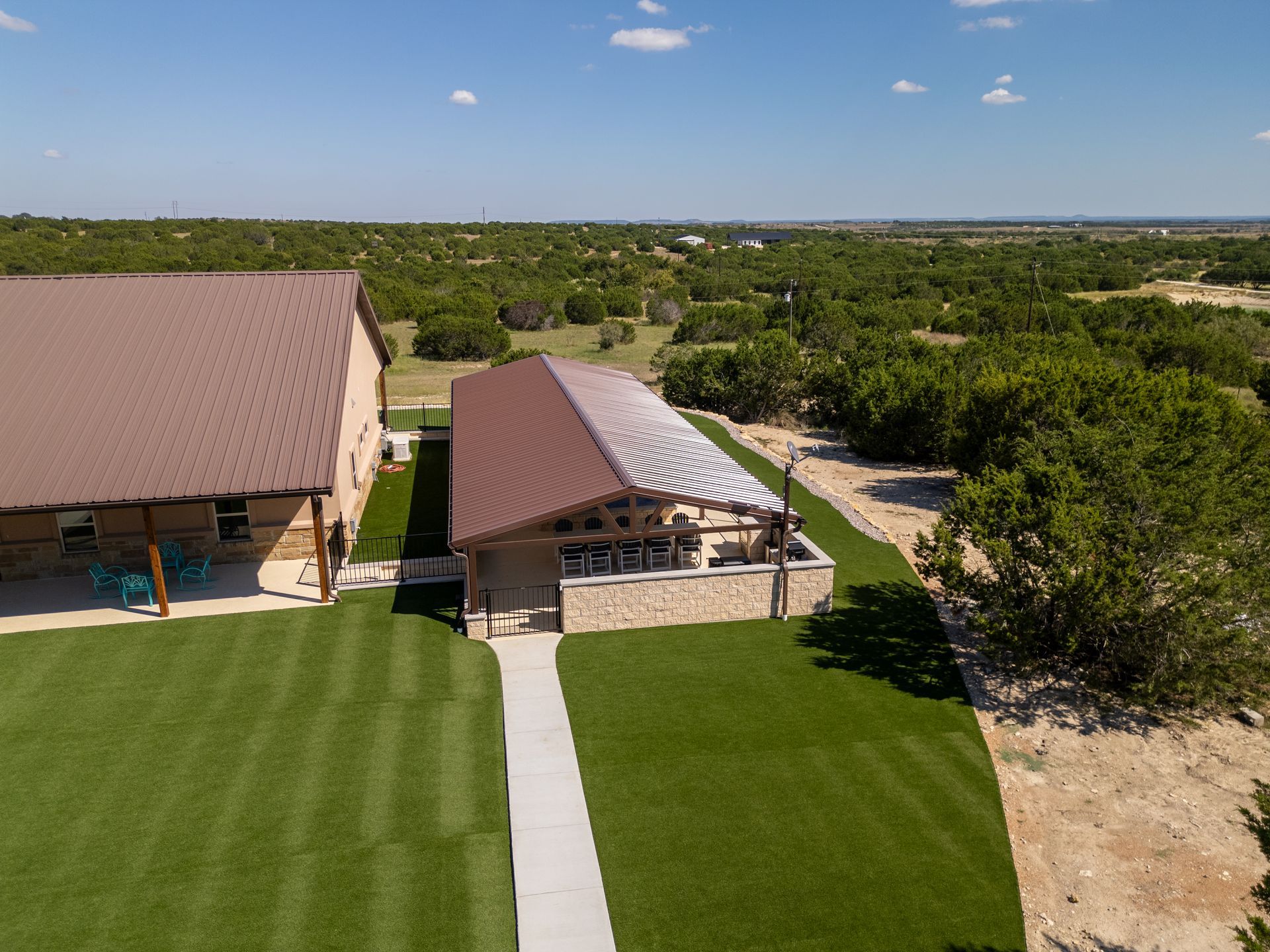 Aerial view of two structures with brown metal roofs surrounded by a lawn and a landscape of green trees under a blue sky.