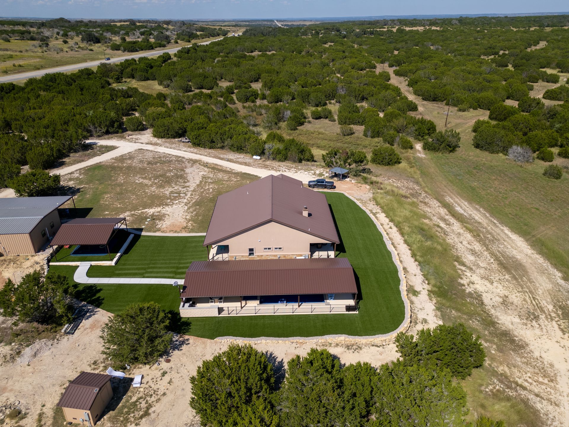Aerial view of a house with a brown metal roof, a green lawn, and a small outbuilding on a rural, wooded property.