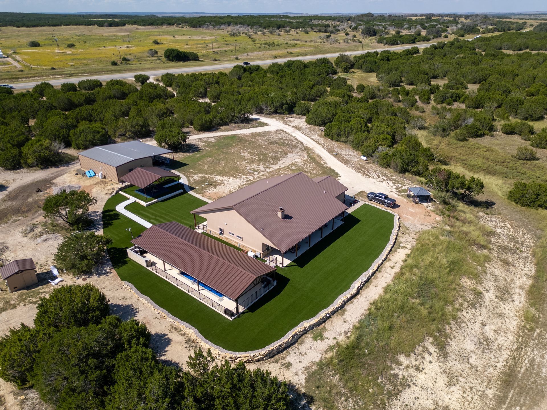 An aerial view of a property featuring several brown-roofed buildings, a green lawn, and a stone retaining wall.