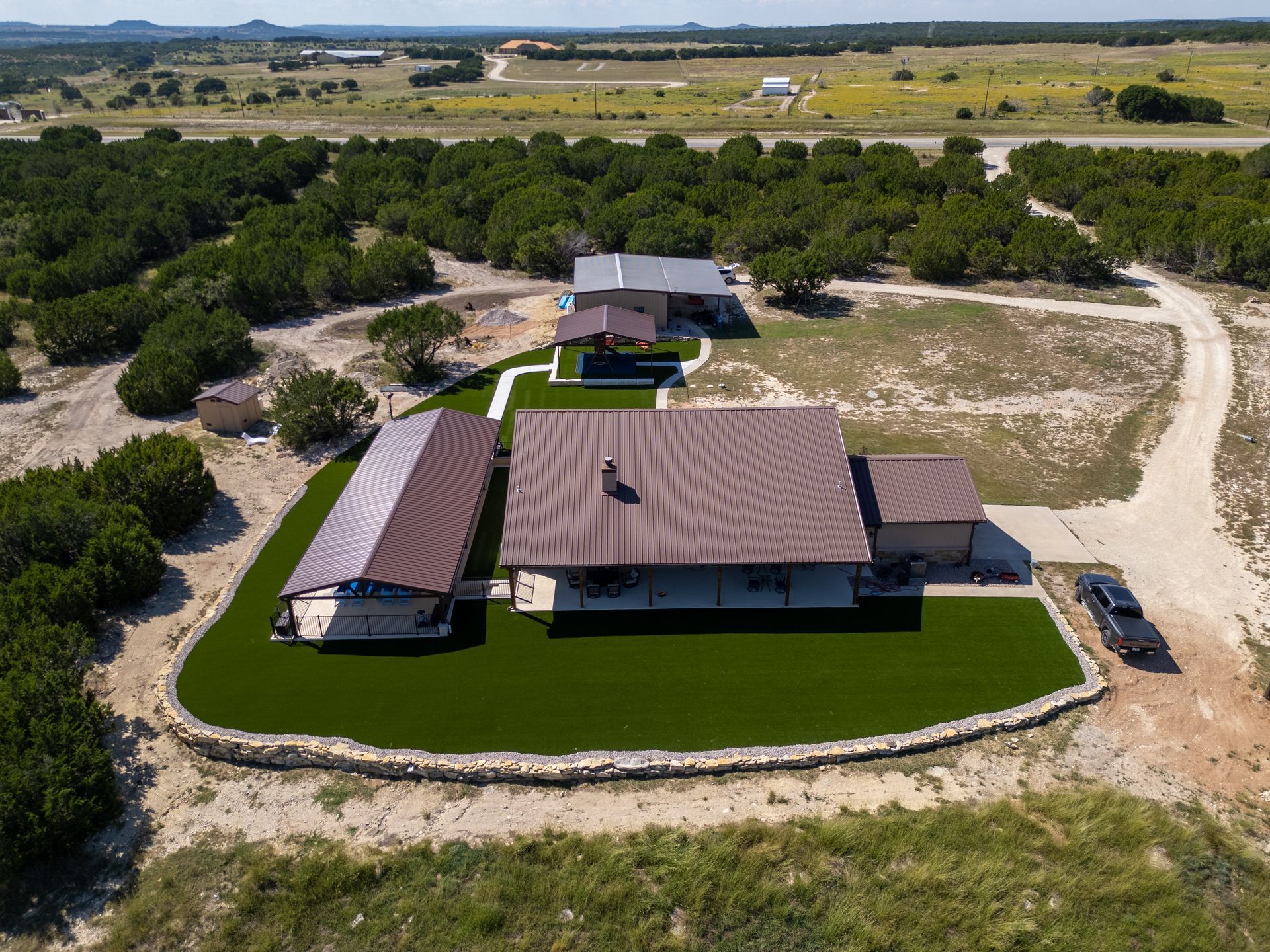 Aerial view of a rural home with a brown metal roof, stone retaining wall, and green lawn, surrounded by wooded terrain.
